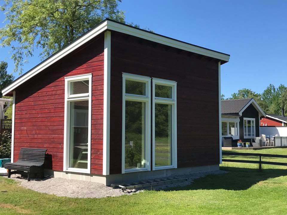Rödmålad komplementbyggnad med pulpettak och stora vita fönster på öländsk tomt